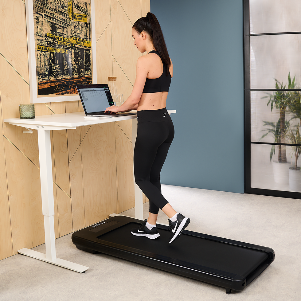 A woman is walking on a treadmill while working on a laptop placed on a standing desk. She is dressed in athletic wear and the setting appears to be a modern home office.